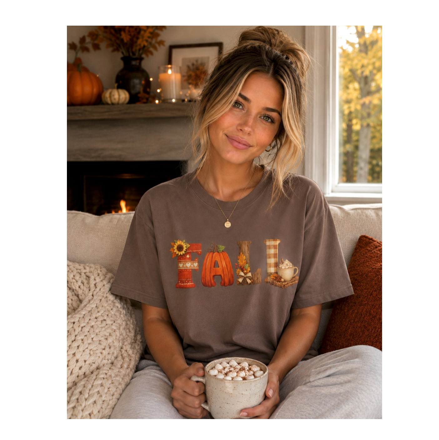 A woman sitting on a couch in a cozy living room setting, wearing a brown t-shirt with a colorful 'FALL' graphic made of autumn-themed icons. She is holding a mug filled with hot chocolate and marshmallows.