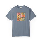 A slate blue short-sleeve t-shirt with a retro-style multicolor graphic on the chest that reads 'IN MY MOM ERA' in wavy, bubble letters.