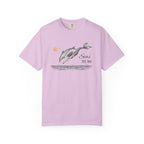 A light purple short-sleeve crewneck t-shirt featuring a graphic print of two dolphins jumping over water with the text 'Seas THE DAY' and a small orange sun.