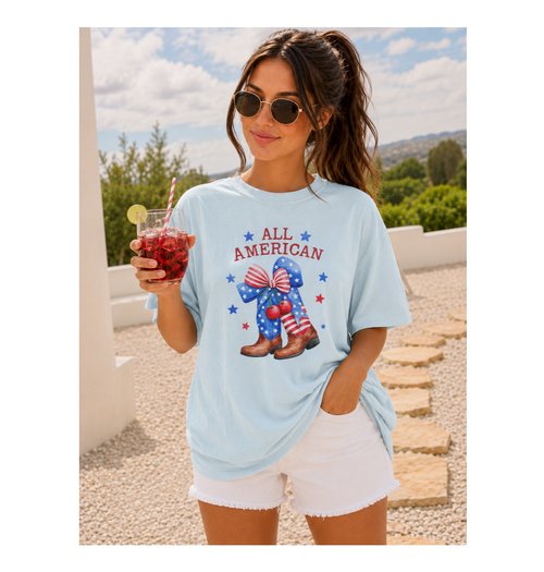 Woman wearing a light blue t-shirt with 'All American' graphic, holding a drink outdoors.