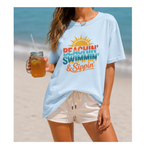 Person wearing a light blue t-shirt with 'Beachin', Swimmin', & Sippin' text on a beach.