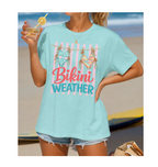 Woman wearing a light blue t-shirt with 'Bikini Weather' text at the beach.