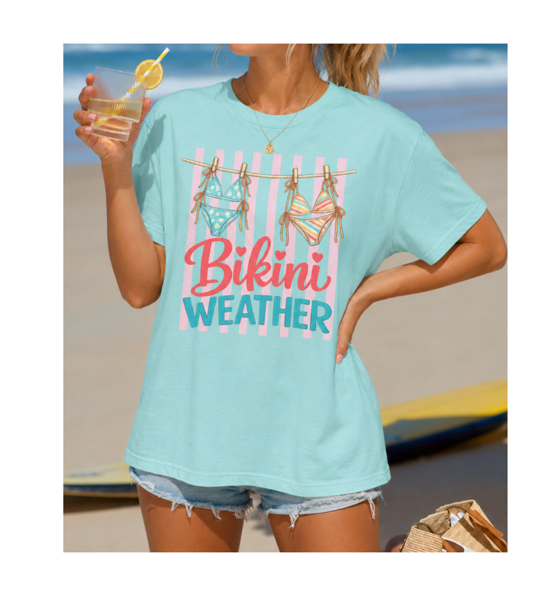 Woman wearing a light blue t-shirt with 'Bikini Weather' text at the beach.