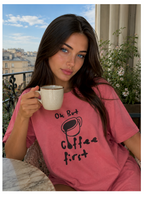 Woman in pink t-shirt with 'Coffee first' text holding a coffee cup outdoors.