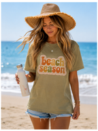Woman on a beach wearing a 'Beach Season' t-shirt with a straw hat and water bottle.