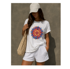 A woman wearing a white crewneck t-shirt with a colorful mandala graphic on the front, paired with white shorts, a beige baseball cap, and carrying a beige crochet shoulder bag against a textured gray wall.