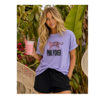 A woman wearing a lavender short-sleeve graphic T-shirt with a pink leopard print and the words 'PINK POWER' in black, paired with black shorts, holding a pink drink in front of a tropical garden background.