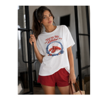 A woman wearing a white graphic t-shirt with a lobster design and the text 'YOU'RE MY Lobster', paired with red shorts, standing in a modern interior setting.