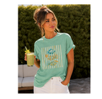 A woman wearing a mint green graphic t-shirt with a striped and starfish design, paired with white pants, holding a drink in front of a hedge background.