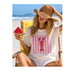 A woman sitting in a beach chair on the sand wears a white T-shirt with a pink striped square and a red lobster graphic, along with a straw sun hat and white shorts. She is holding a slice of watermelon.
