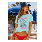 A woman wearing a light aqua graphic t-shirt with 'SUMMER VIBES' and beach chair illustrations in pink and navy, paired with red bikini bottoms and a navy cap, standing in a sunny beach setting with blue umbrellas.