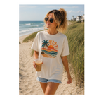 A woman walking on a sandy beach wearing a cream-colored graphic t-shirt with a sunset and palm tree design, paired with denim shorts and sunglasses.