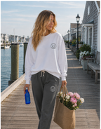 A woman walking on a wooden dock by the water wearing a white crewneck sweatshirt and dark gray jogger sweatpants, both featuring a small circular logo on the chest and thigh. She is carrying a blue water bottle and a jute bag of pink and white flowers.