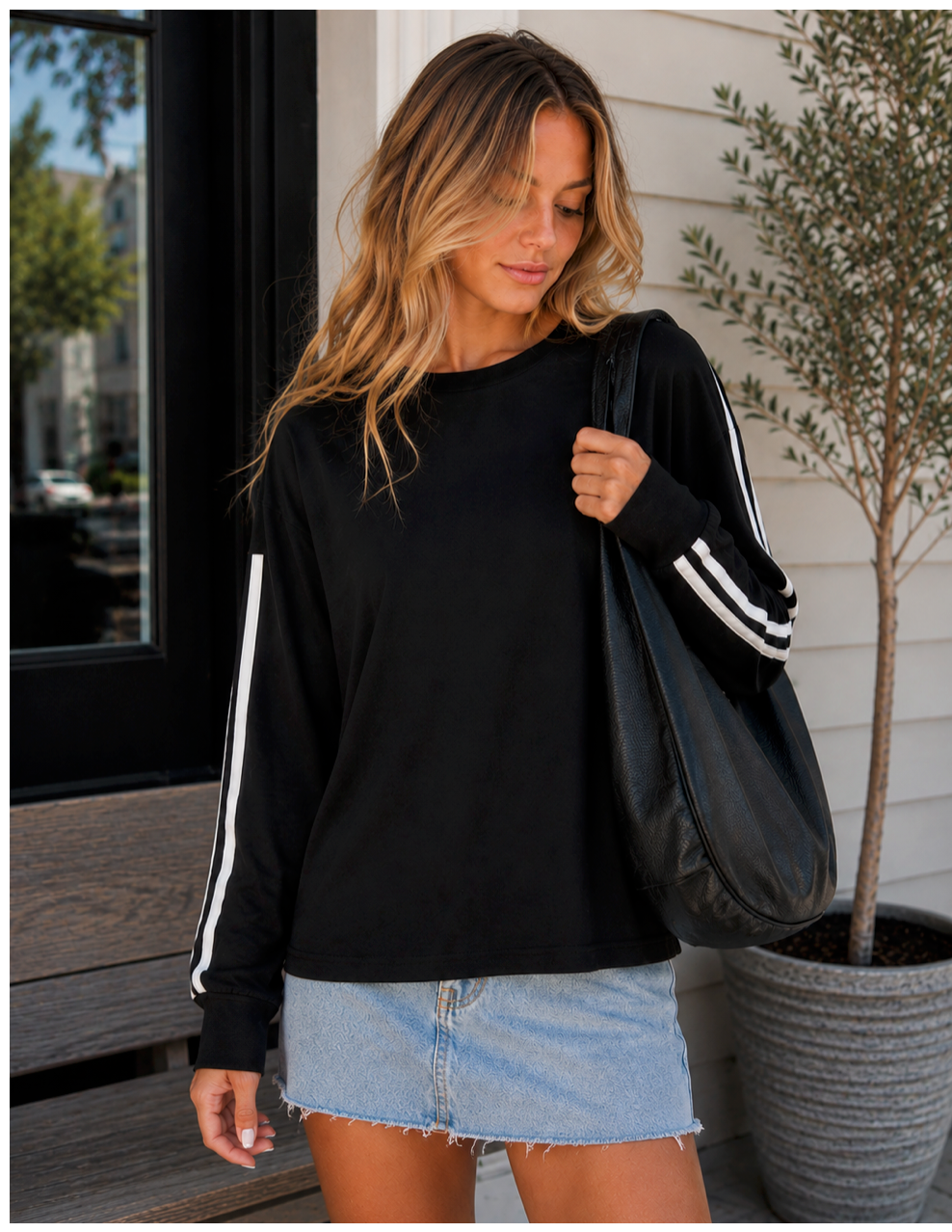 A woman wearing a black long-sleeve sweatshirt with white stripes down the sleeves, paired with a light blue denim mini skirt and carrying a black tote bag.