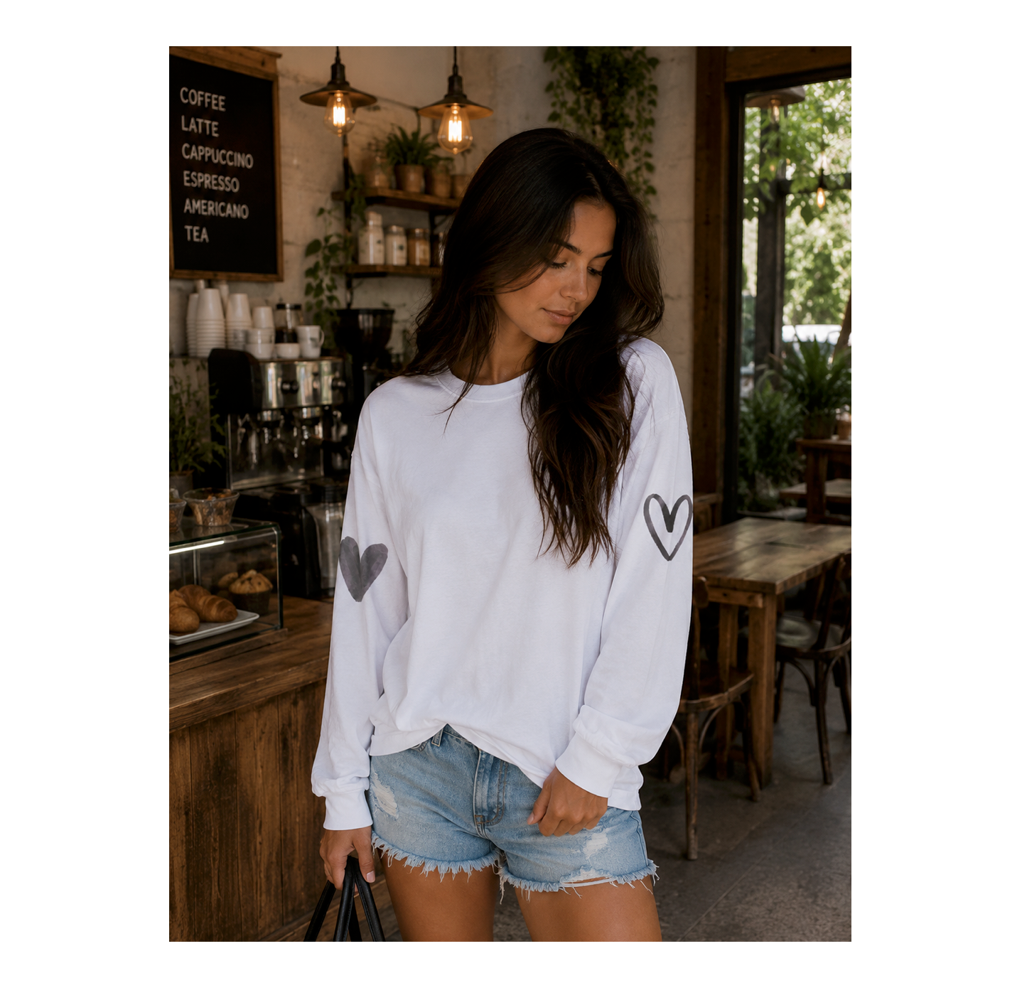 A woman wearing a white long-sleeve sweatshirt with black heart graphics on the sleeves, paired with denim shorts, standing in a cozy cafe environment.