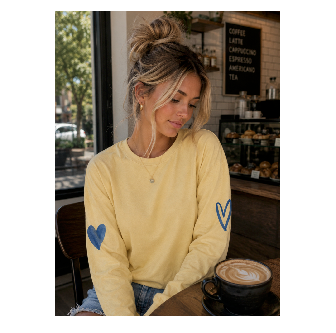 A woman sitting at a cafe table wearing a pale yellow long-sleeve shirt with blue heart designs on both sleeves, paired with blue jeans and a black coffee cup in front of her.
