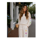 A woman stands on a city street wearing a cream-colored sweatshirt and matching sweatpants, both decorated with colorful embroidered flowers, hearts, and stars. She is holding an iced coffee in front of a storefront.