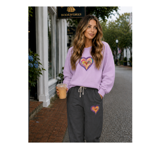 A woman standing on a street wearing a lavender sweatshirt and dark gray sweatpants, both featuring a colorful heart graphic on the front. She is holding an iced coffee in front of a storefront with a 'Bookworks' sign.