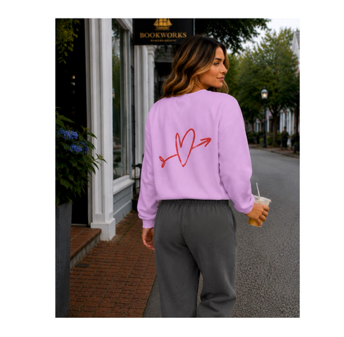 A woman stands on a city street wearing a lavender sweatshirt with a red heart-and-arrow graphic on the back, paired with dark gray sweatpants, holding an iced coffee.