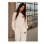 A woman modeling a matching cream sweatshirt and sweatpants lounge set with embroidered heart details, standing in an industrial loft setting with a large window and concrete columns.