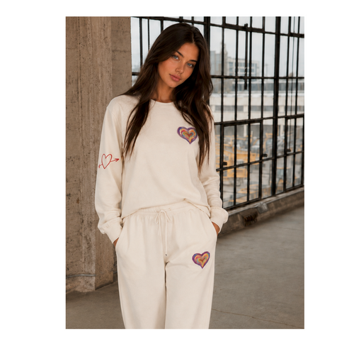 A woman modeling a matching cream sweatshirt and sweatpants lounge set with embroidered heart details, standing in an industrial loft setting with a large window and concrete columns.