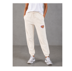 A person wearing cream-colored jogger sweatpants with a small colorful heart patch on the left thigh, paired with a white t-shirt and silver sneakers, standing against a gray studio background.