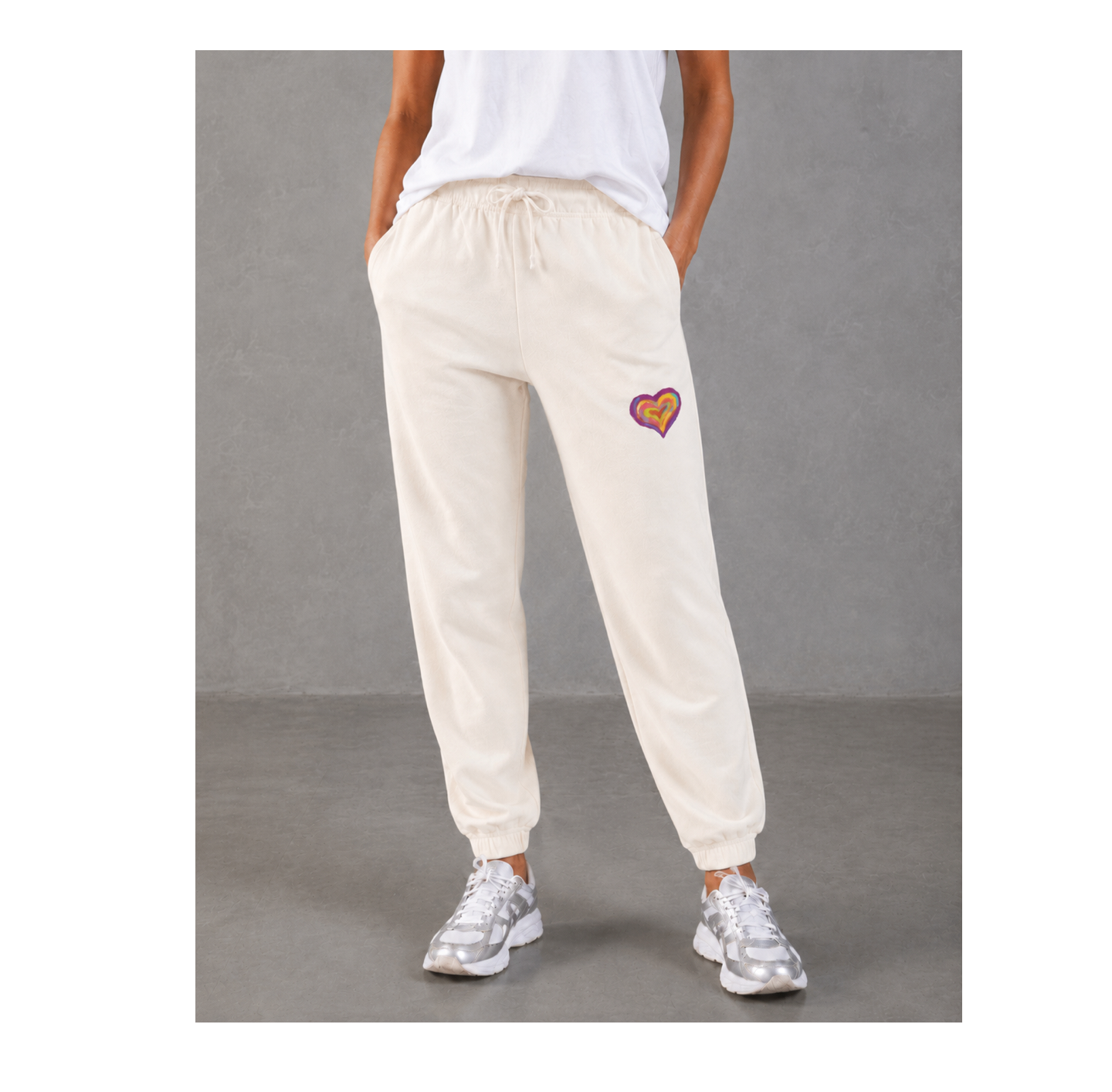 A person wearing cream-colored jogger sweatpants with a small colorful heart patch on the left thigh, paired with a white t-shirt and silver sneakers, standing against a gray studio background.