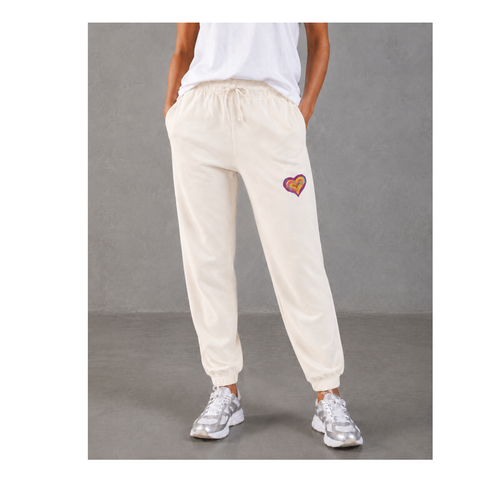 A person wearing cream-colored jogger sweatpants with a small colorful heart patch on the left thigh, paired with a white t-shirt and silver sneakers, standing against a gray studio background.