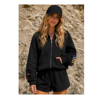 A woman wearing a black zip-up hoodie with colorful striped sleeve accents and matching black shorts, standing outdoors against a rocky, sandy background.