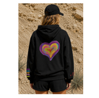 A person shown from behind wearing a black hoodie with a large colorful heart graphic on the back and a black cap, standing outdoors against a sandy desert background.