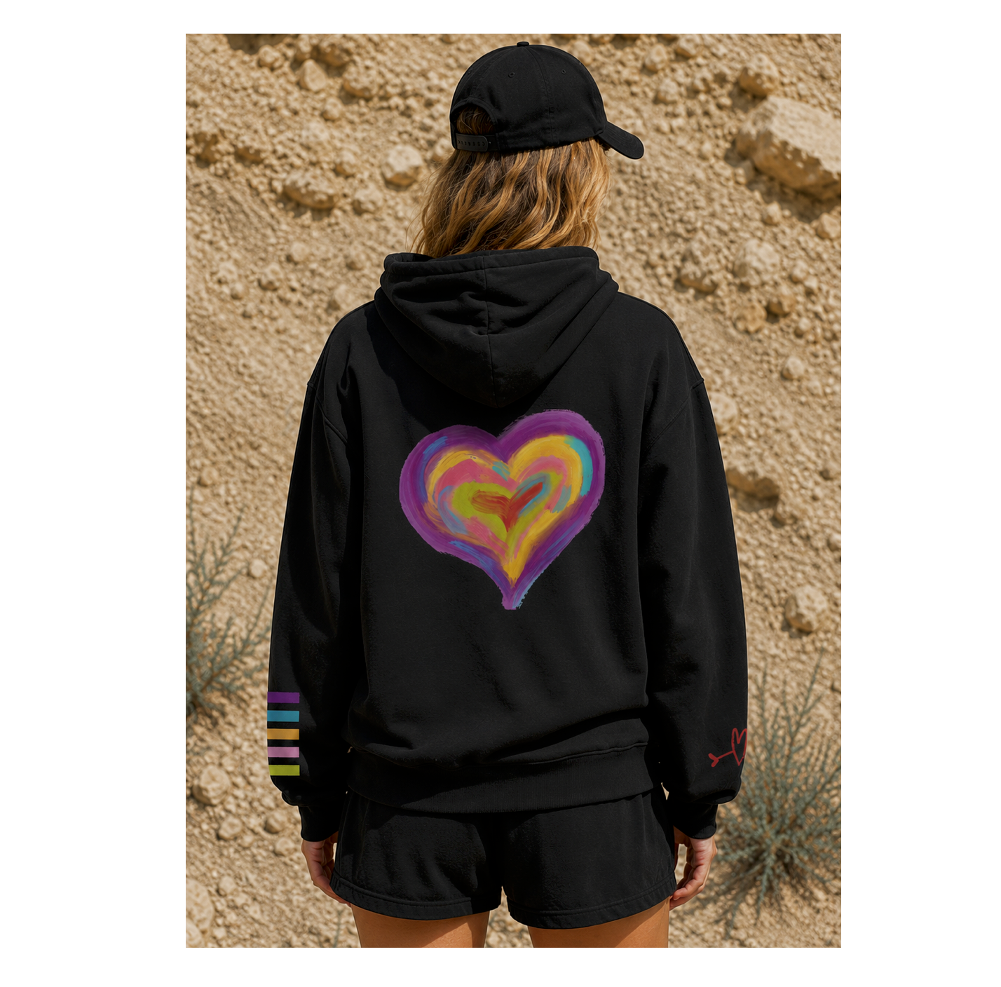 A person shown from behind wearing a black hoodie with a large colorful heart graphic on the back and a black cap, standing outdoors against a sandy desert background.