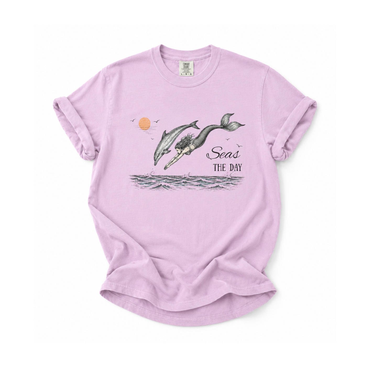 A light purple short-sleeve t-shirt with a graphic of two whales jumping out of the water, a small orange sun, and the text 'Seas THE DAY'.