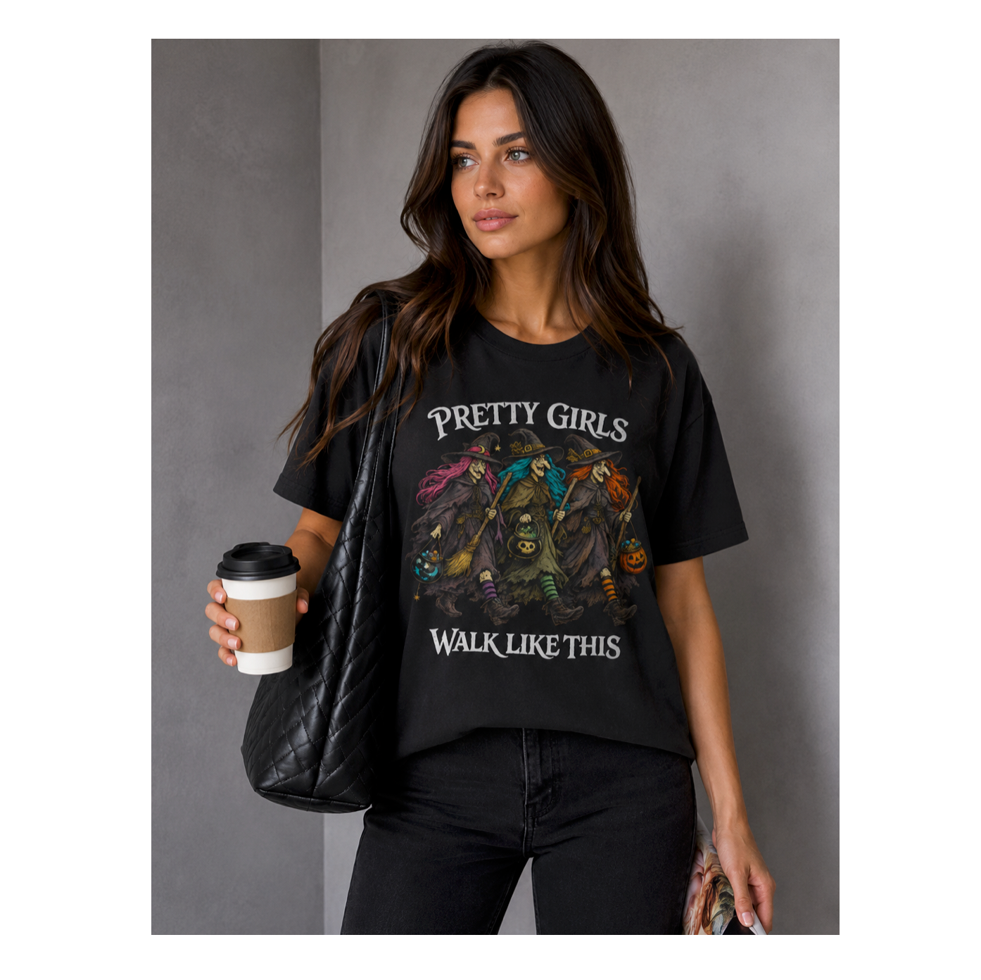 A woman modeling a black graphic t-shirt with the text 'PRETTY GIRLS WALK LIKE THIS' and a colorful illustration of three girls. She is holding a black quilted shoulder bag and a coffee cup against a gray background.