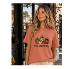 A woman wearing a dusty rose short-sleeve graphic t-shirt with a fall-themed pumpkin print and the text 'WILD ABOUT FALL' on the front, set against a blurred storefront background.