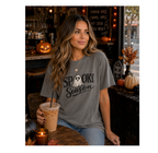 A woman wearing a dark gray graphic t-shirt that reads 'Spooky Season' with a white ghost design, sitting in a cozy, dimly lit cafe with pumpkins and autumn decorations.