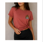 A woman wearing a dusty rose short-sleeve t-shirt with rolled cuffs and a side knot, featuring a small leopard-print chest graphic with a bow. She is paired with black bottoms against a neutral gray background.