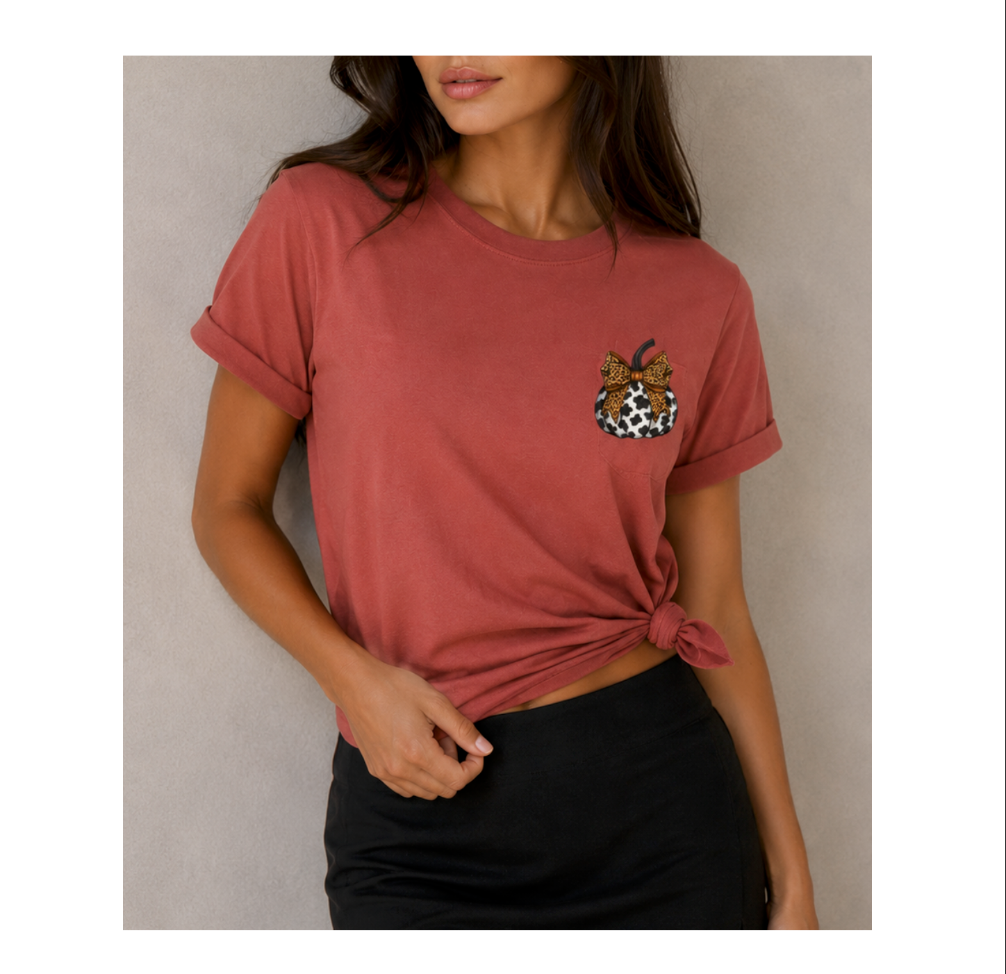 A woman wearing a dusty rose short-sleeve t-shirt with rolled cuffs and a side knot, featuring a small leopard-print chest graphic with a bow. She is paired with black bottoms against a neutral gray background.