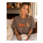 A woman sitting on a couch in a cozy living room setting, wearing a brown t-shirt with a 'Fall' graphic print. She is holding a mug filled with hot chocolate and marshmallows.