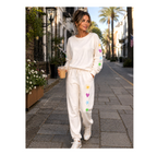 A woman walking down a city street wearing a cream-colored sweatshirt and sweatpants set with colorful embroidered hearts and stars. She is holding an iced coffee.