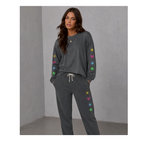 A woman modeling a dark gray lounge set consisting of a long-sleeve sweatshirt and matching sweatpants, decorated with colorful heart and star motifs on the sleeves and legs, set against a gray textured wall.