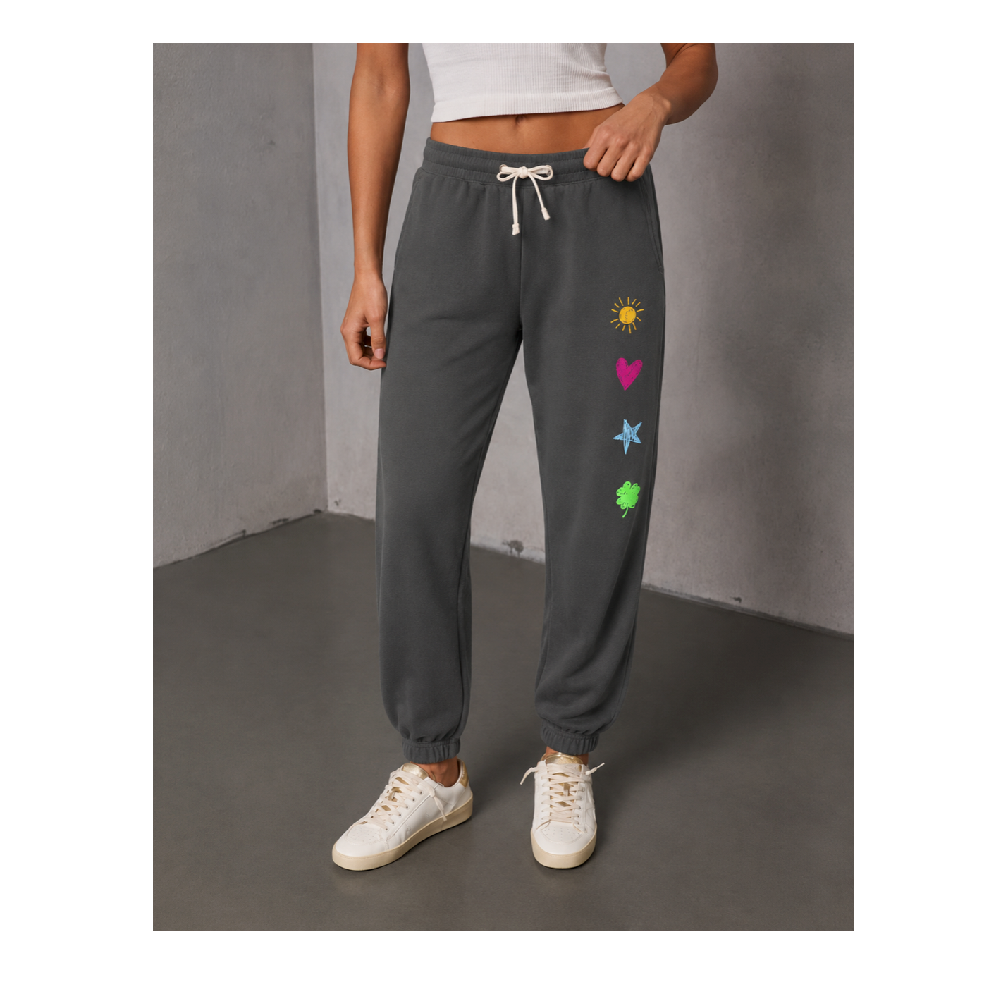 A person wearing dark gray jogger sweatpants with a white drawstring and colorful heart, star, and clover graphics on the left leg, paired with white sneakers in a gray studio setting.