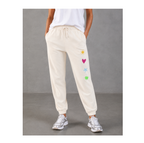 A person wearing cream-colored jogger sweatpants with a drawstring waist and colorful embroidered sun, heart, star, and flower motifs on the left leg, paired with a white top and silver sneakers against a gray studio background.