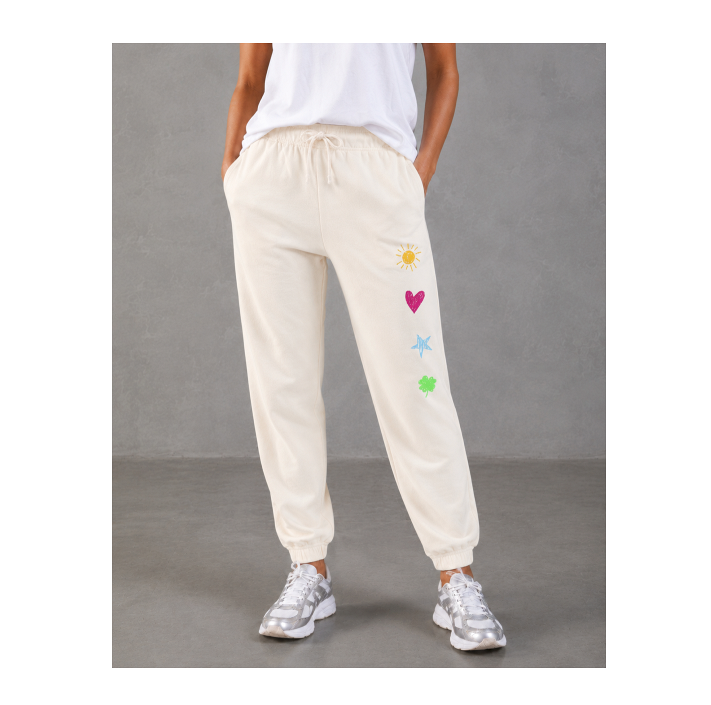 A person wearing cream-colored jogger sweatpants with a drawstring waist and colorful embroidered sun, heart, star, and flower motifs on the left leg, paired with a white top and silver sneakers against a gray studio background.