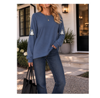 A woman wearing a slate blue long-sleeve sweatshirt with white ghost graphics on the sleeves, paired with dark blue jeans, standing on a house porch and holding a black tote bag.