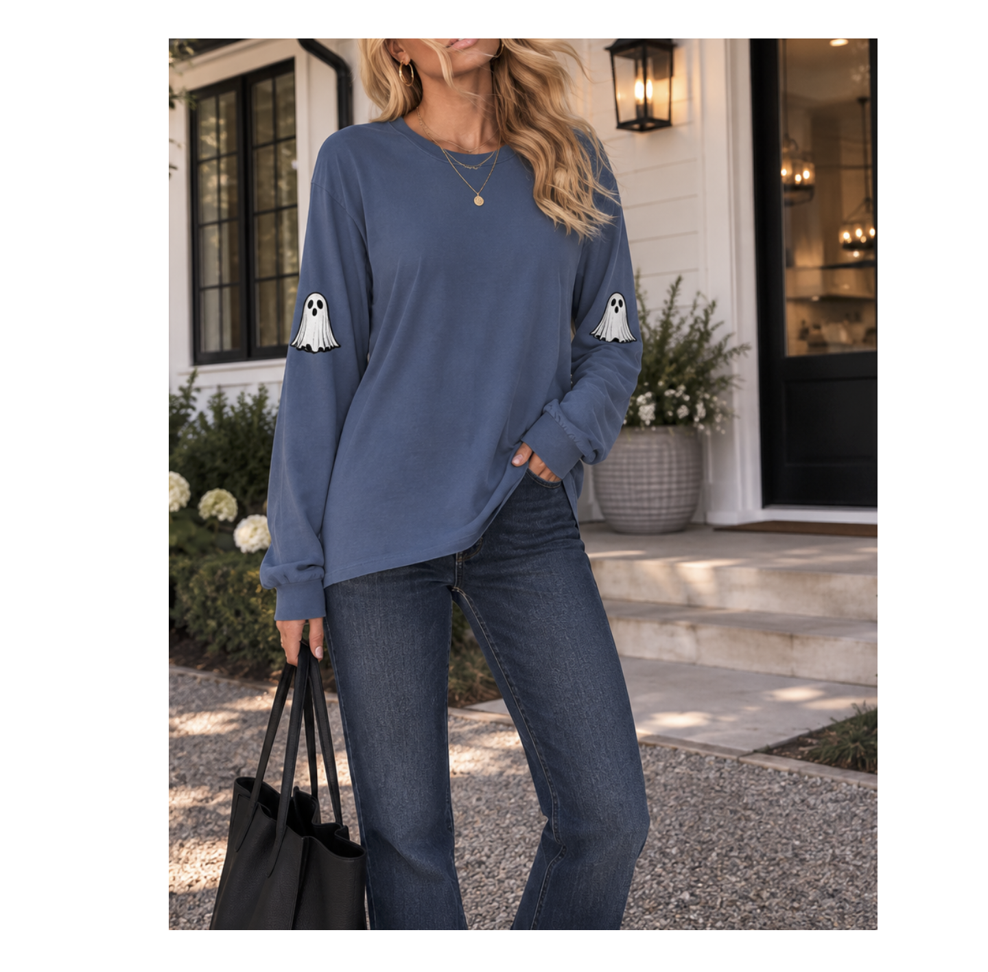 A woman wearing a slate blue long-sleeve sweatshirt with white ghost graphics on the sleeves, paired with dark blue jeans, standing on a house porch and holding a black tote bag.