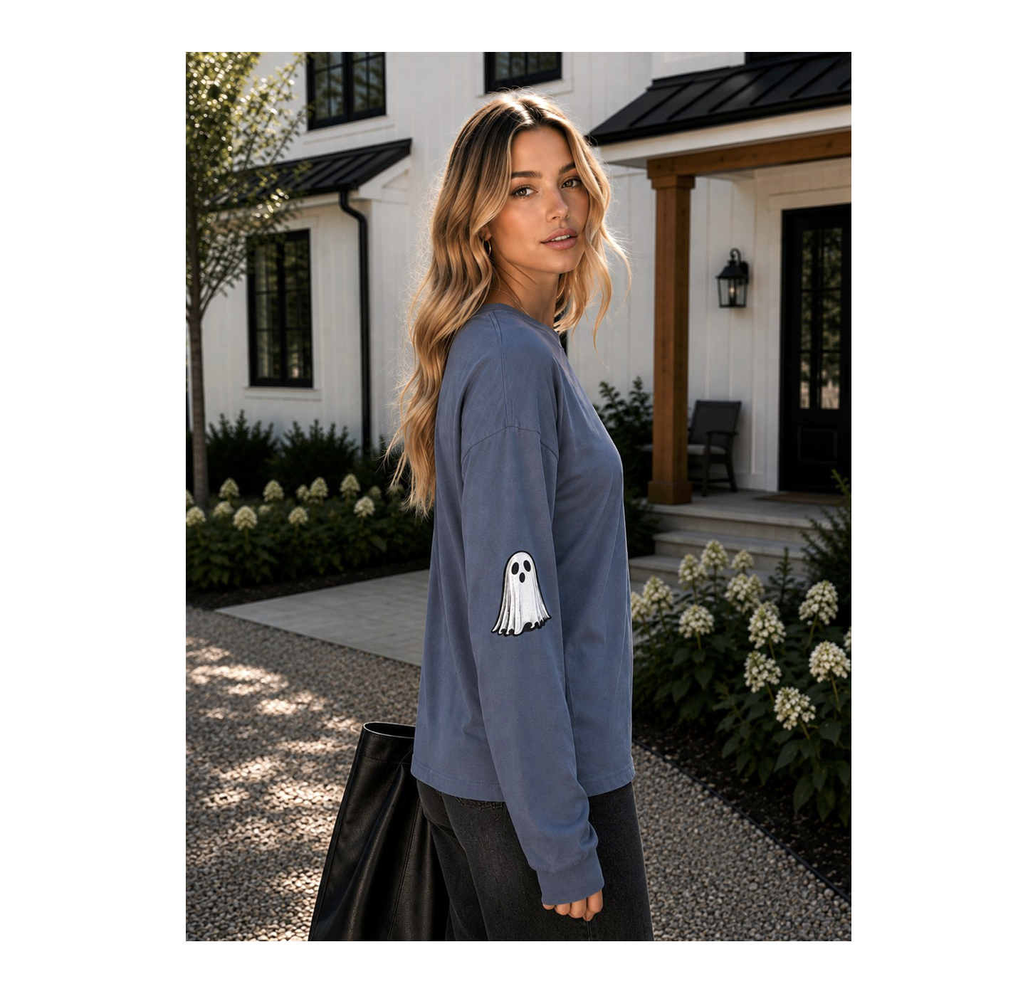 A woman stands outdoors in front of a white house with a porch, wearing a slate blue long-sleeve shirt with a white ghost graphic on the sleeve, paired with dark pants and carrying a black tote bag.