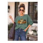 A woman models a sage green graphic sweatshirt with a fall harvest design featuring pumpkins, a leopard print pattern, and the text 'WILD ABOUT FALL'. She is wearing blue jeans and carrying a brown leather tote bag, standing in front of a rustic porch with pumpkins.