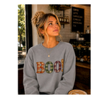 A woman wearing a light gray crewneck sweatshirt with the word 'BOO!' in a colorful plaid pattern, sitting inside a cozy cafe.