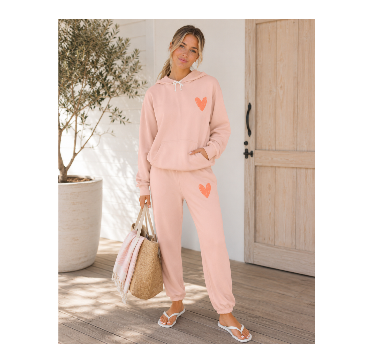 A woman modeling a light pink two-piece loungewear set with coral heart graphics on the hoodie and sweatpants, standing on a wooden deck next to a potted tree and holding a straw tote bag.