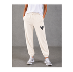 A model from the waist down wearing off-white jogger sweatpants with a black heart patch on the left thigh, paired with a white t-shirt and silver sneakers, set against a gray concrete background.